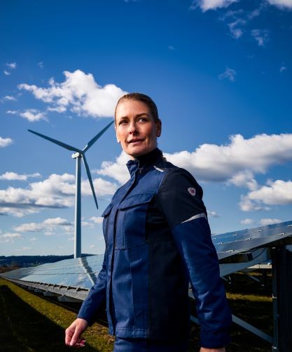 Eine Frau trägt Multinorm Schutzkleidung. Im Hintergrund ist eine Windkraftanlage und Solaranlagen zu sehen.